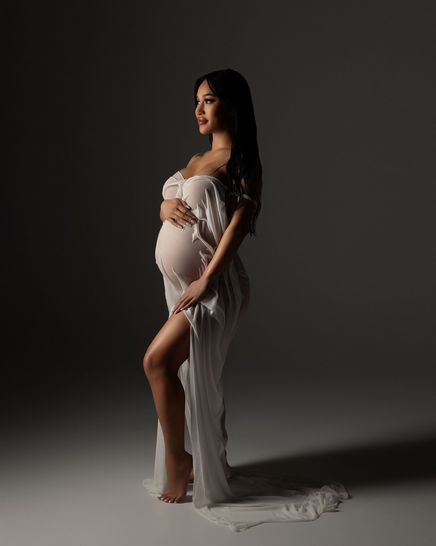 Artistic maternity portrait showing an elegant silhouette in flowing white fabric against a dark studio backdrop.