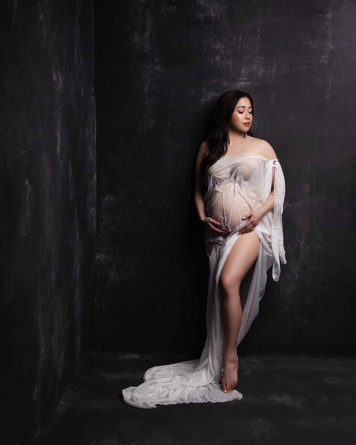 A maternity photoshoot featuring an elegant white lace gown against a dark backdrop in dramatic lighting.