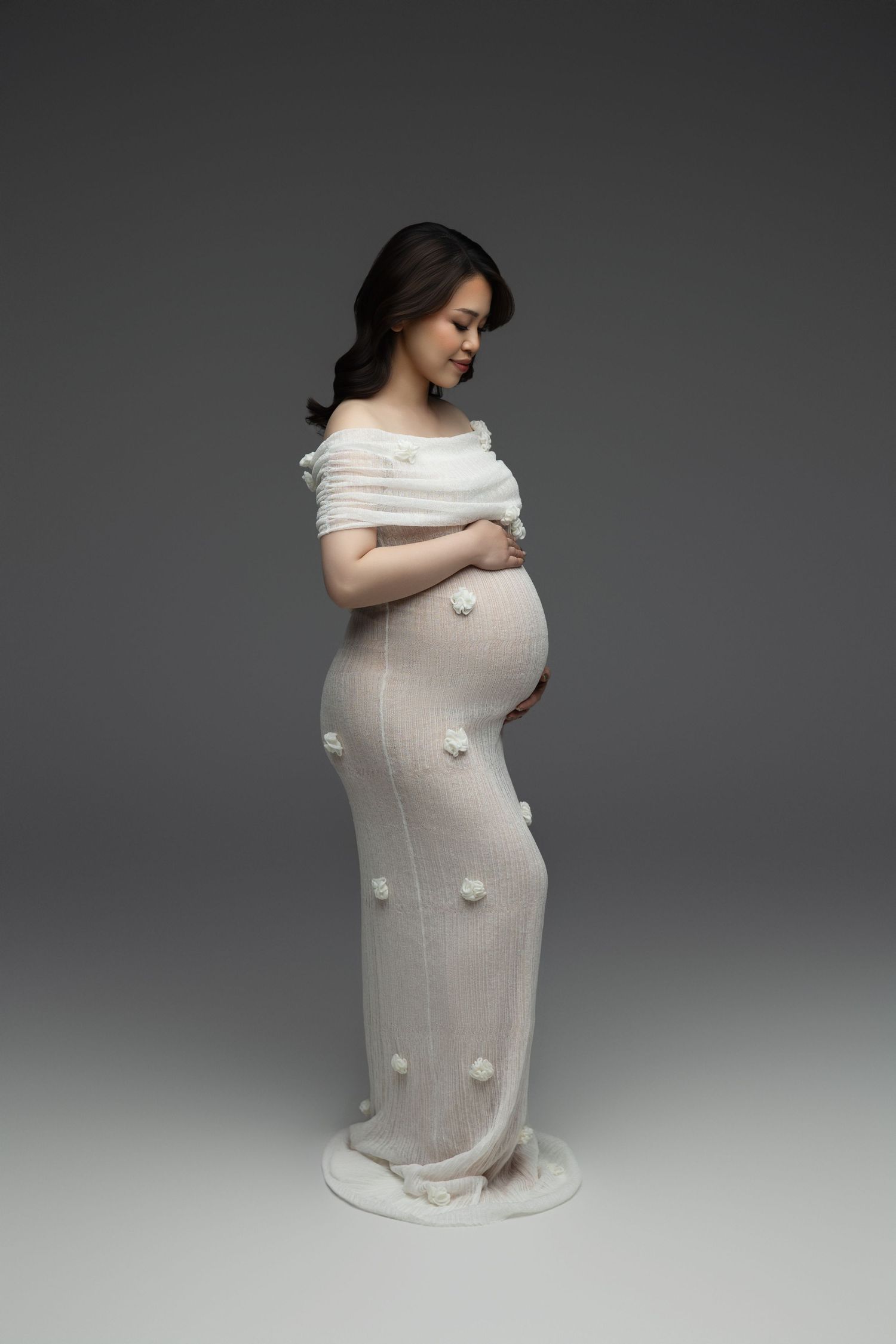 Elegant maternity portrait showing a figure in off-shoulder white dress with floral details standing against dark backdrop.