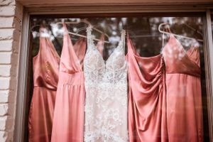 Wedding dress and coral bridesmaid dresses hanging in a row against a window.
