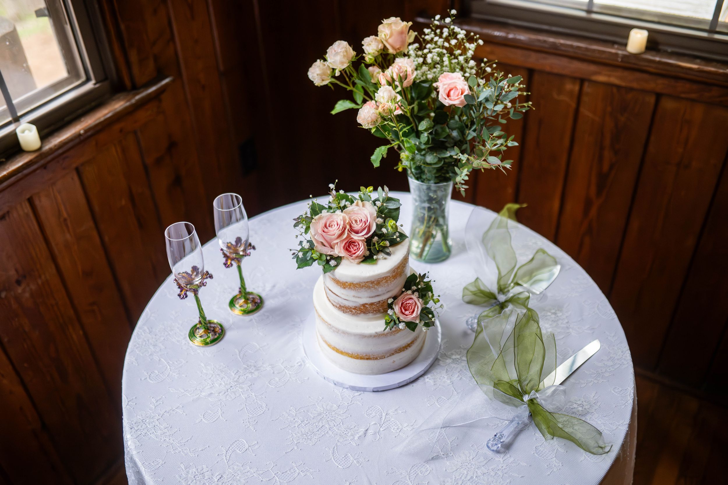 A simple white wedding cake decorated with fresh pink flowers and greenery sits elegantly on a round table.