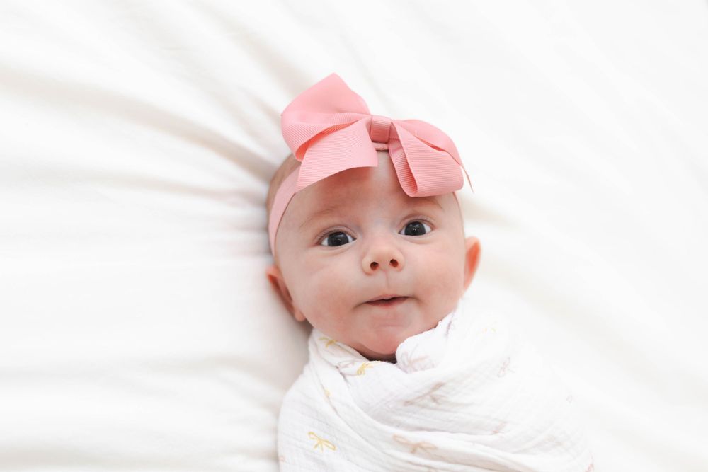 Baby wearing pink bow headband lying on white bedding.