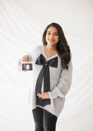 Expectant mother in grey sweater holding ultrasound photo against white backdrop.