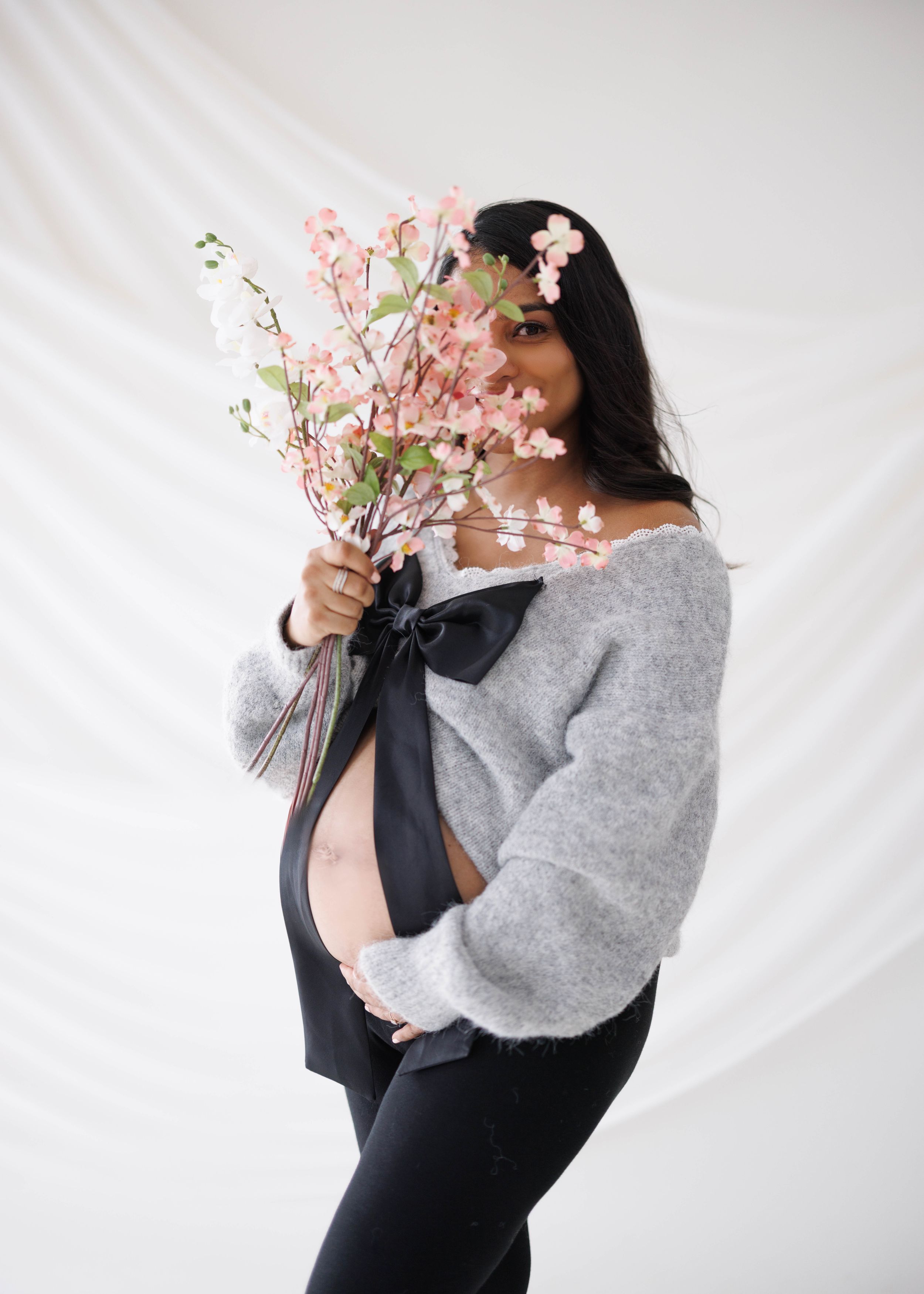 Maternity photo with grey sweater and pink cherry blossoms against white backdrop.