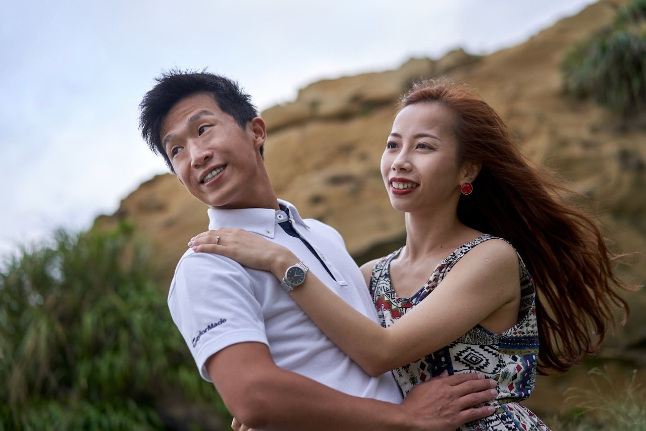 Happy couple embracing and laughing together against rocky cliffside background with flowing brown hair in wind.