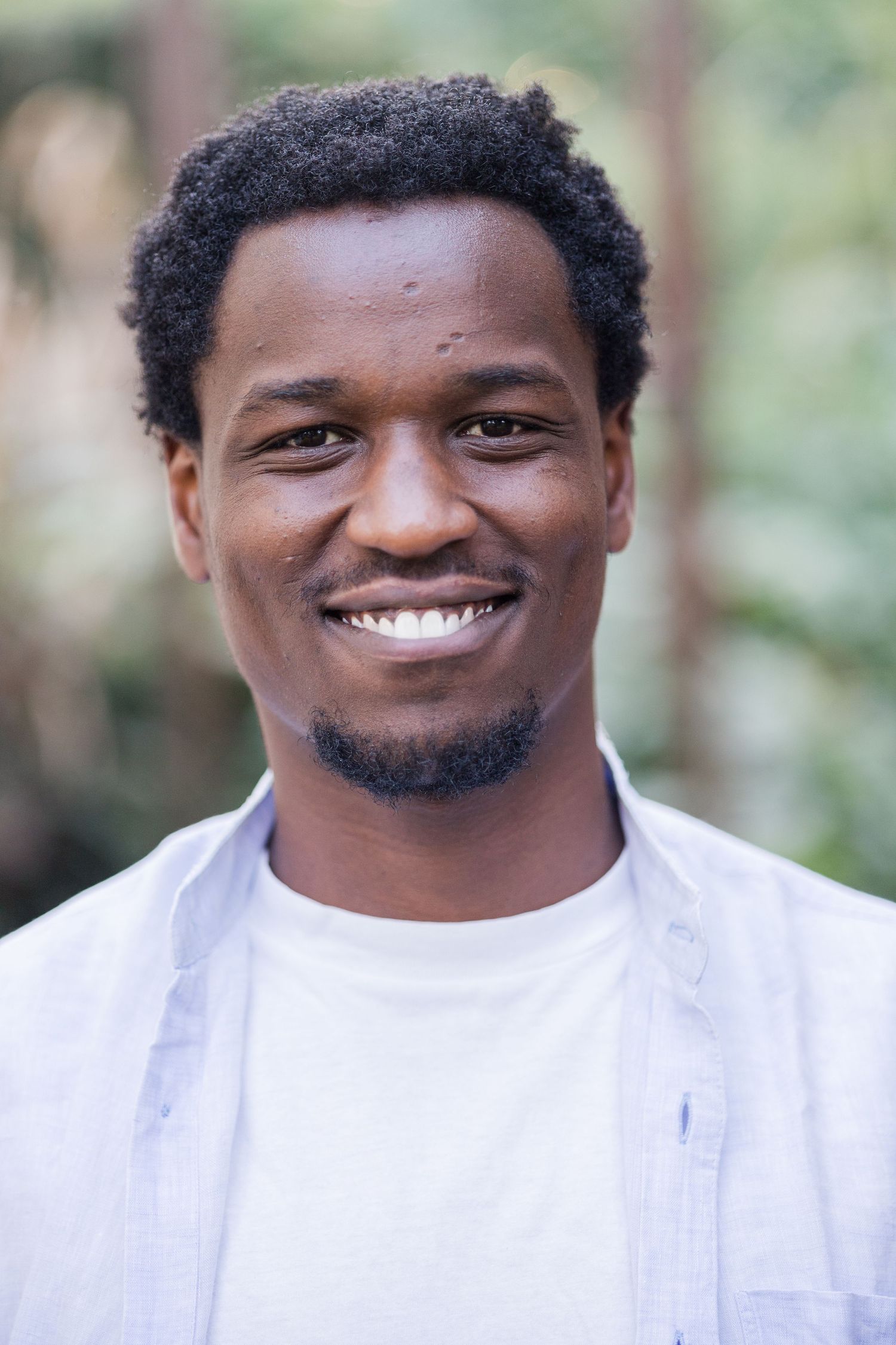 Photo of Ikigu George, smiling at the camera. He has an afro. He's wearing a white t-shirt and light blue shirt.