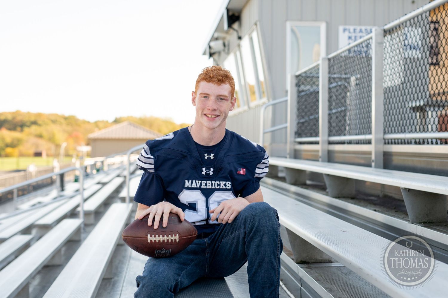 Athlete in navy blue Mavericks jersey poses with football on metal bleachers.