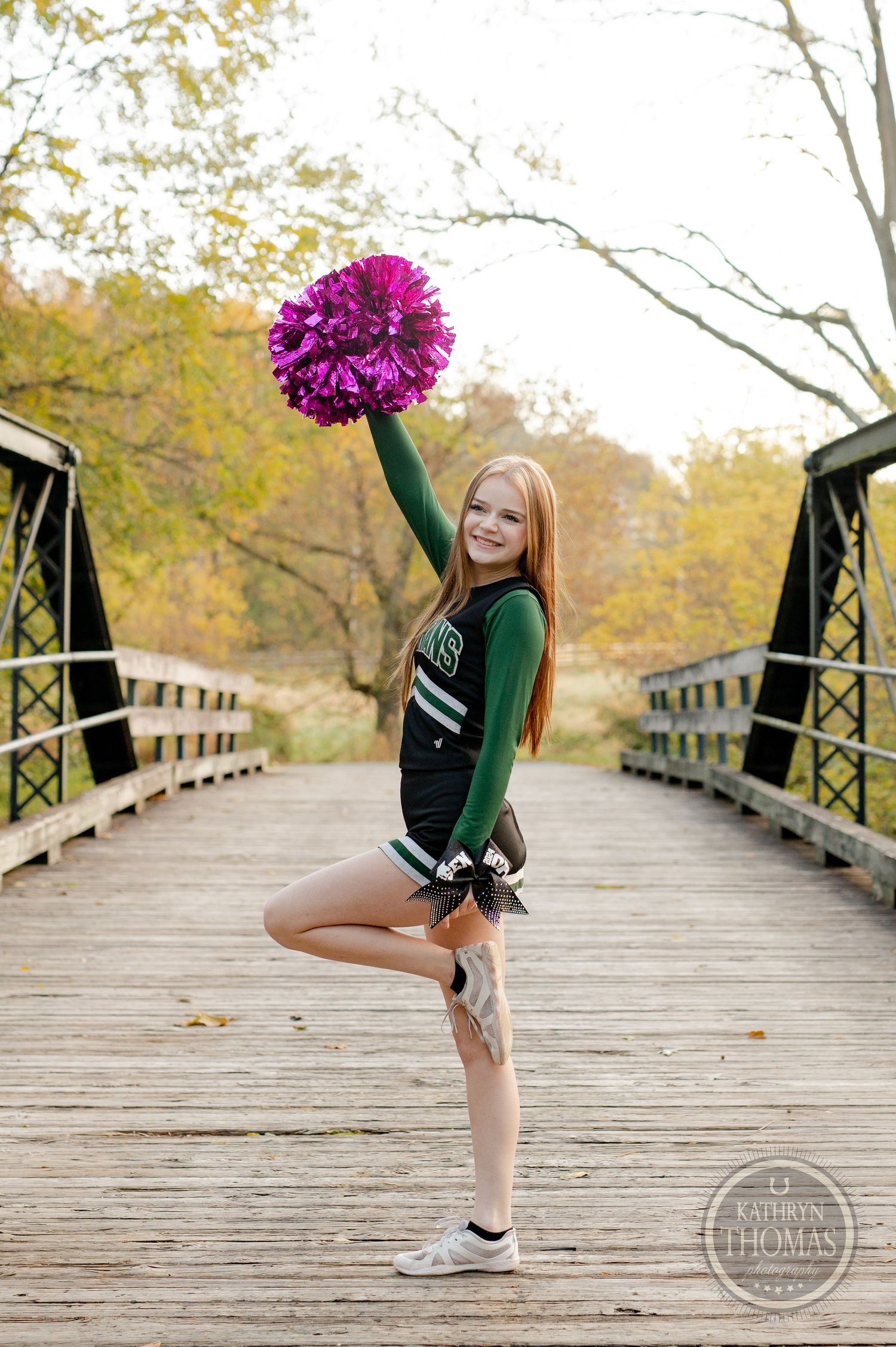 Cheerleader strikes dynamic pose with pink pom pom raised on wooden bridge during autumn.