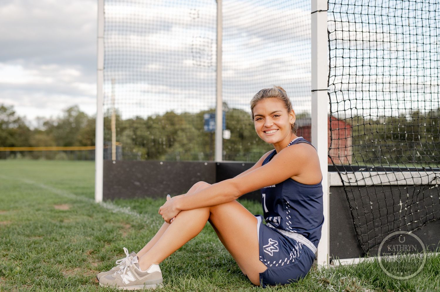 Field hockey player sits against goal post wearing navy uniform on sunny day.