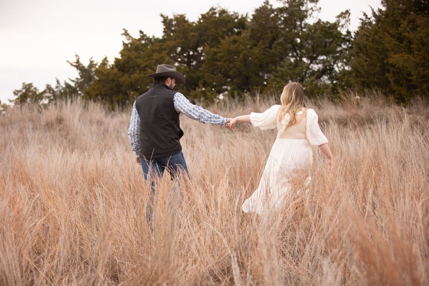 Couple holding hands while walking through tall golden grass in a romantic outdoor setting at sunset.