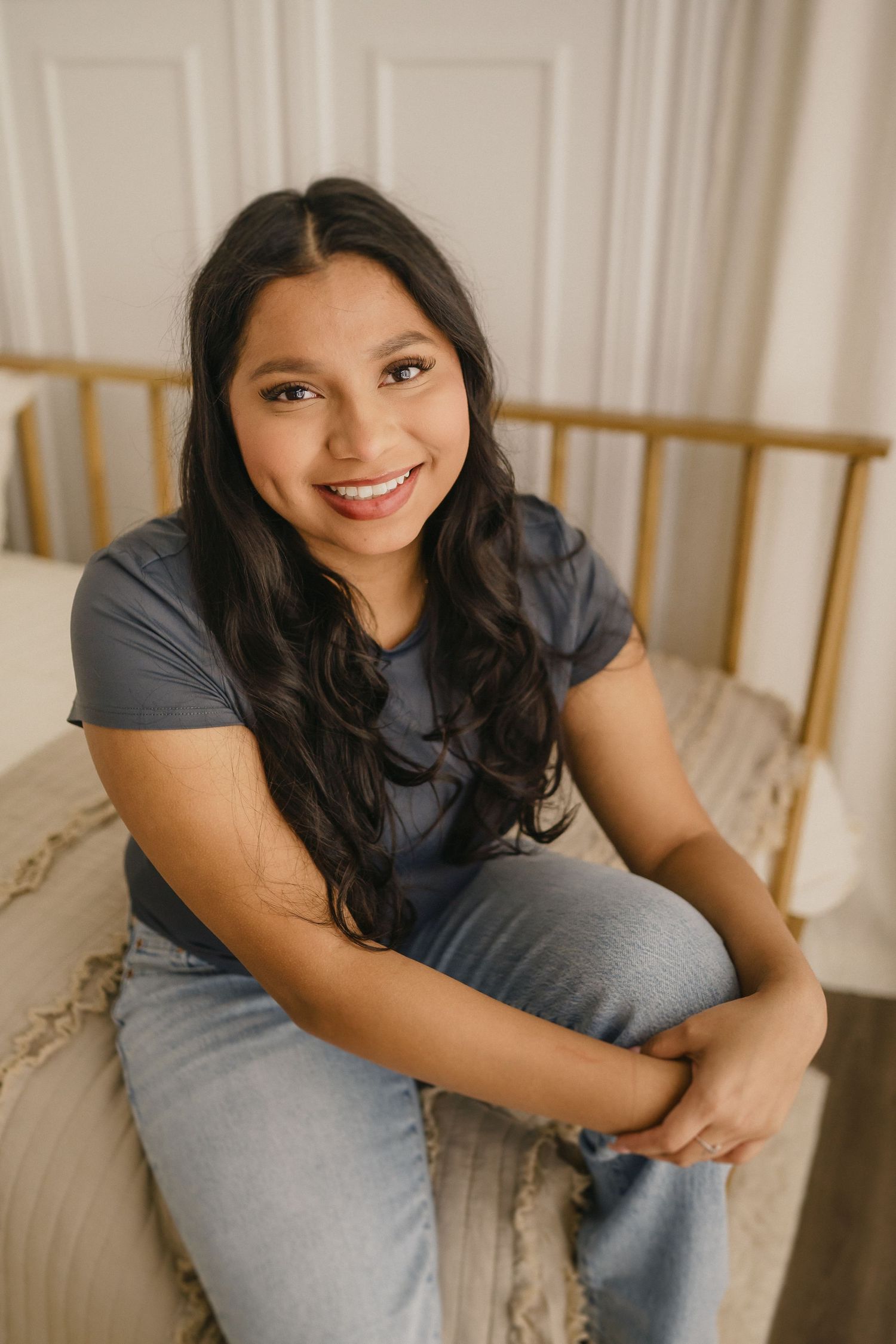 A young woman in casual attire sits on a bed with gold frame, wearing gray shirt and jeans in warm indoor lighting.