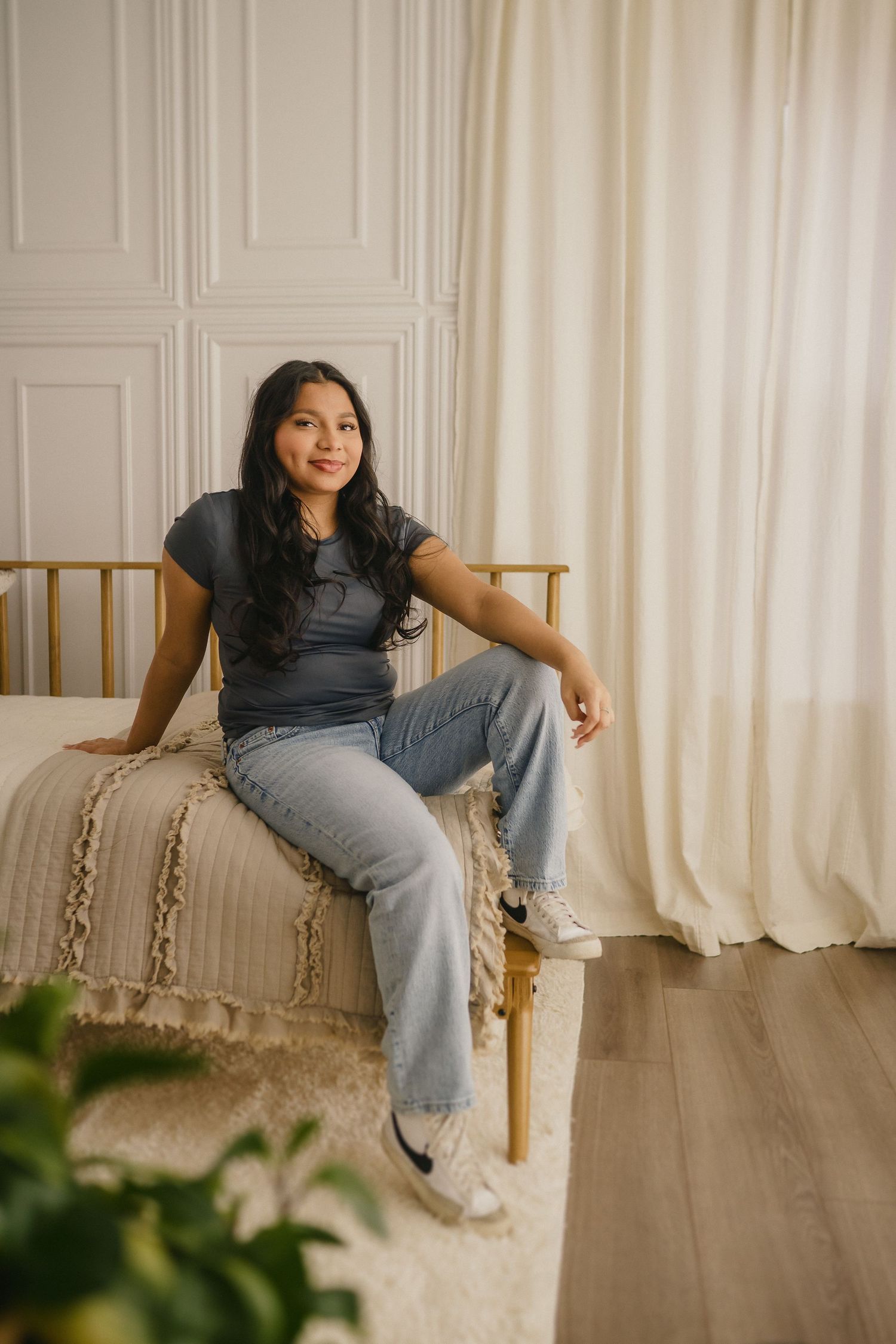 Someone sits casually on a wooden bench wearing a gray t-shirt and jeans in a bright room with white walls and curtains.