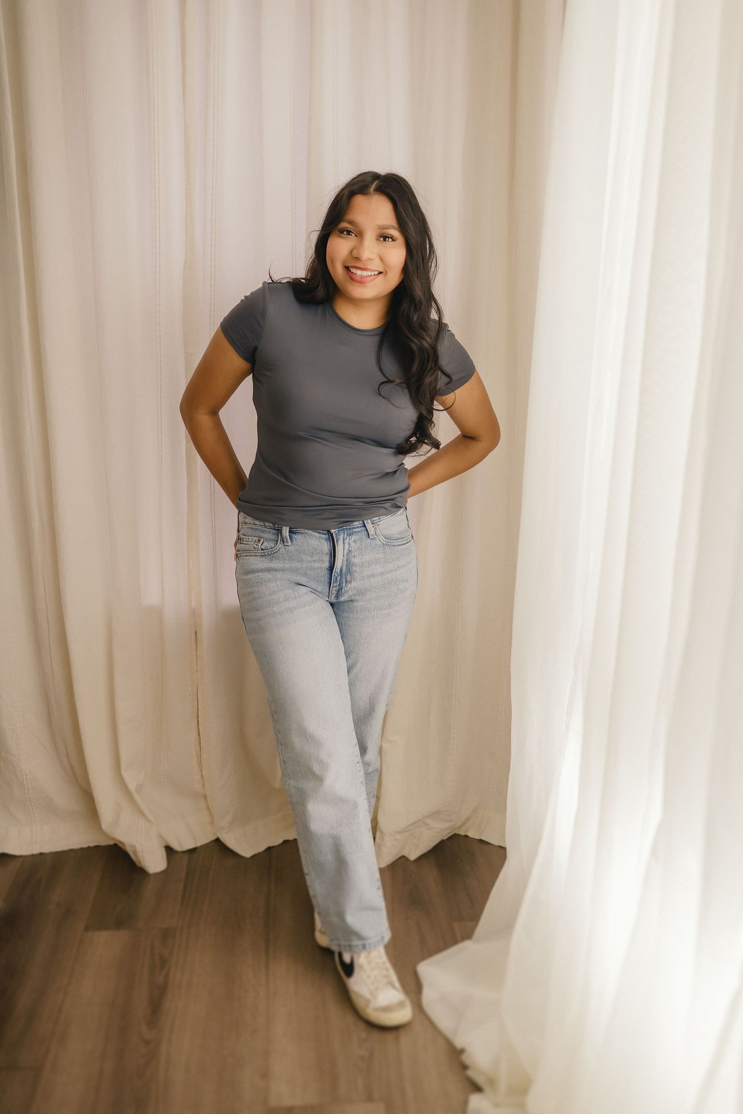 Person in casual outfit with grey t-shirt and light wash jeans poses against white curtain backdrop.