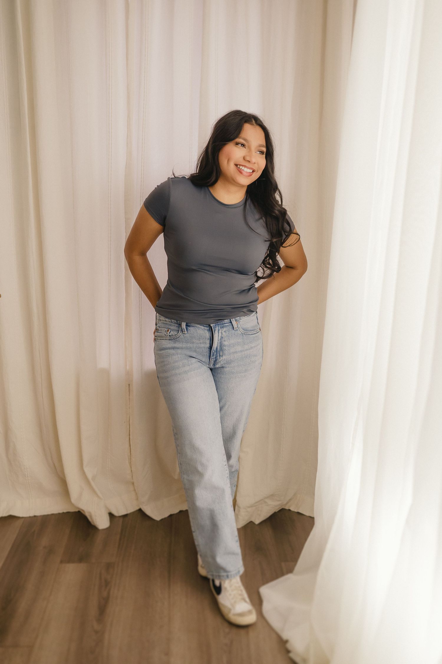 Woman in gray t-shirt and light jeans posing against white curtains on hardwood floor.