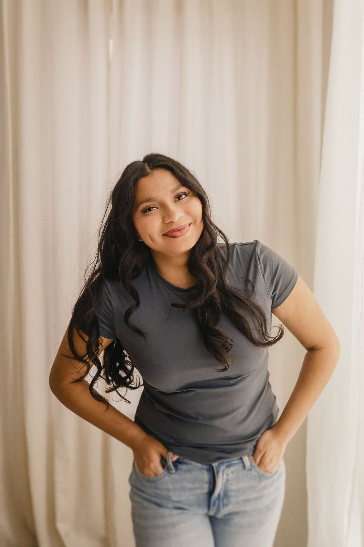 A woman in a gray t-shirt and jeans poses casually against white curtains in a naturally lit interior setting.
