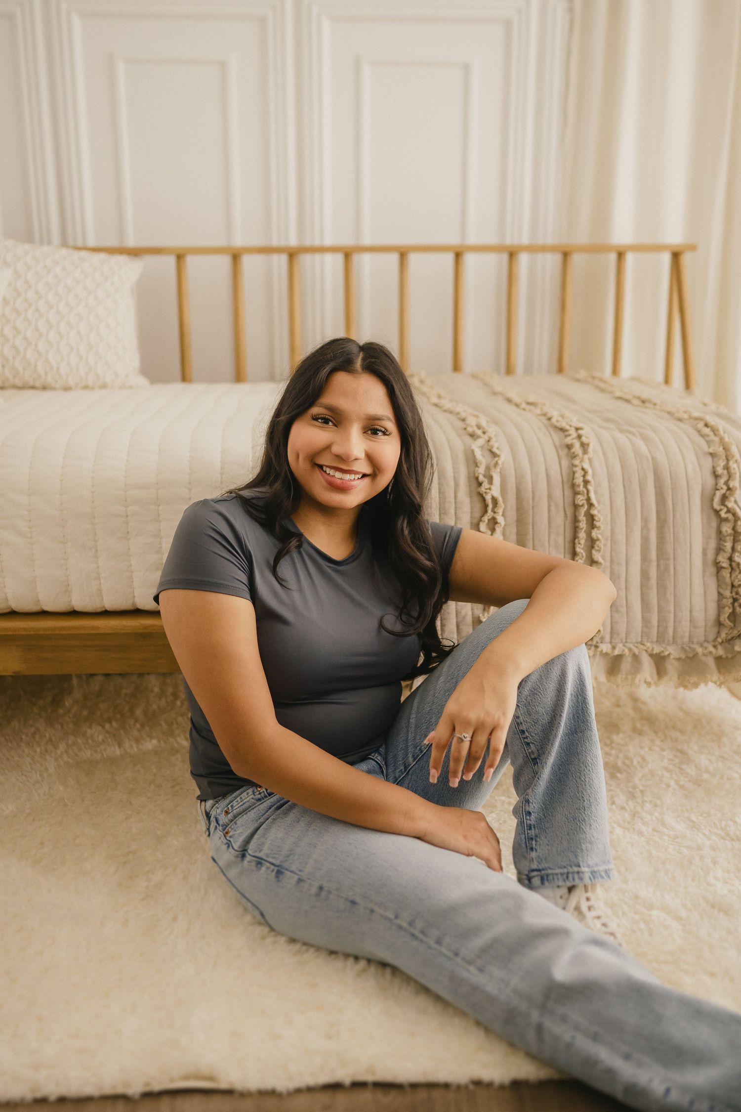 Someone sits casually on a carpeted floor by a bed with brass frame, wearing a gray t-shirt and light wash jeans.