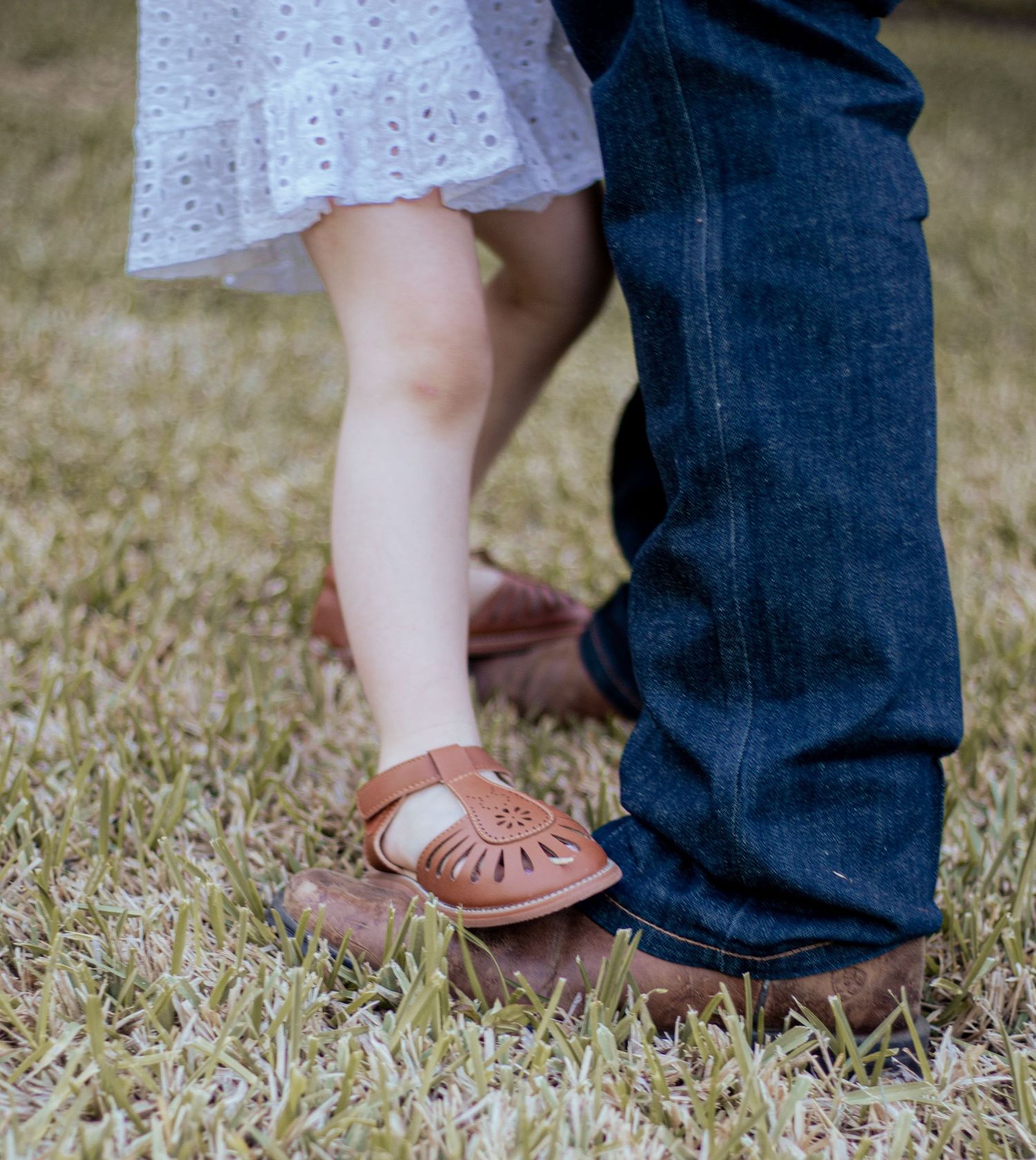 Person wearing a dainty white eyelet dress embraces someone in blue jeans and brown boots in grass.
