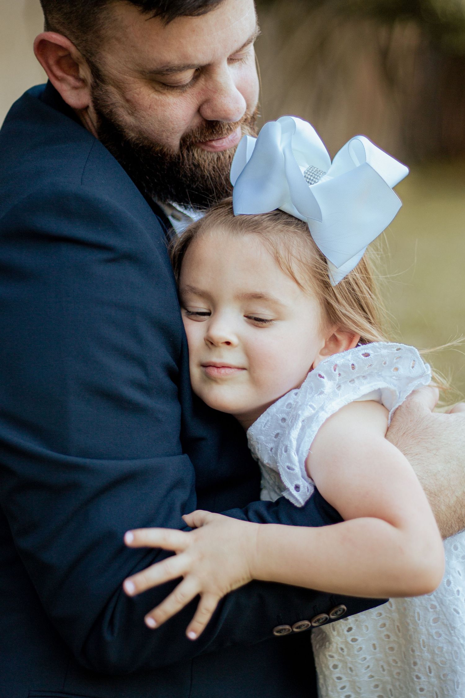 A bearded person in a navy suit embraces a small child wearing a white dress and bow during a tender moment outdoors.