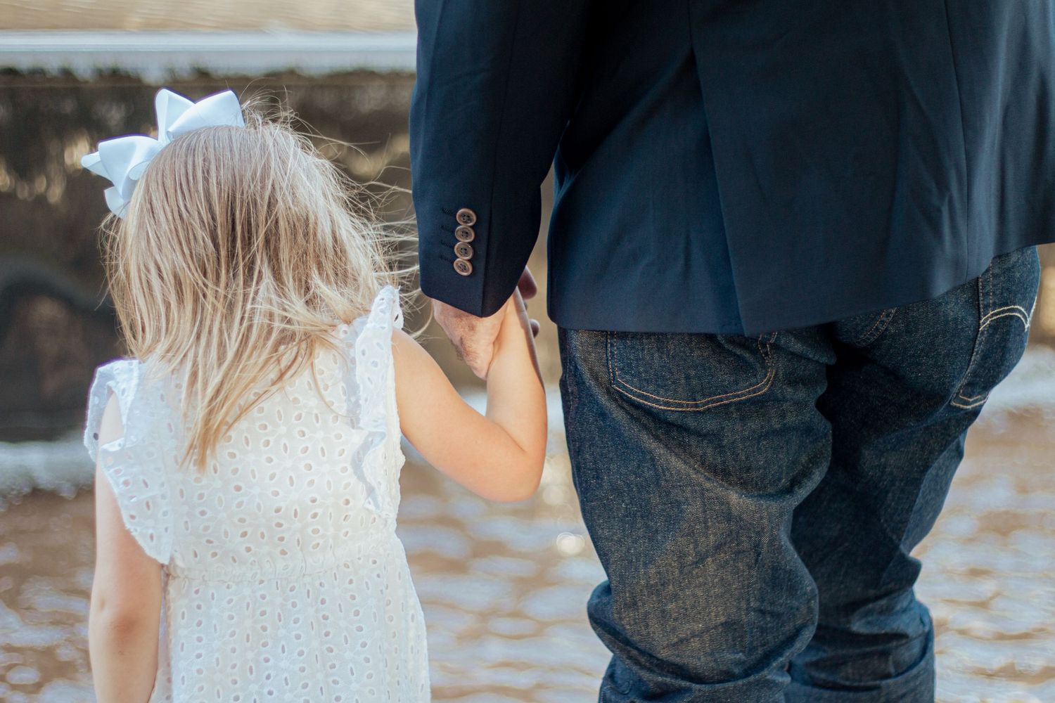 A child in a white lace dress holds hands with an adult wearing jeans while walking on sandy ground.