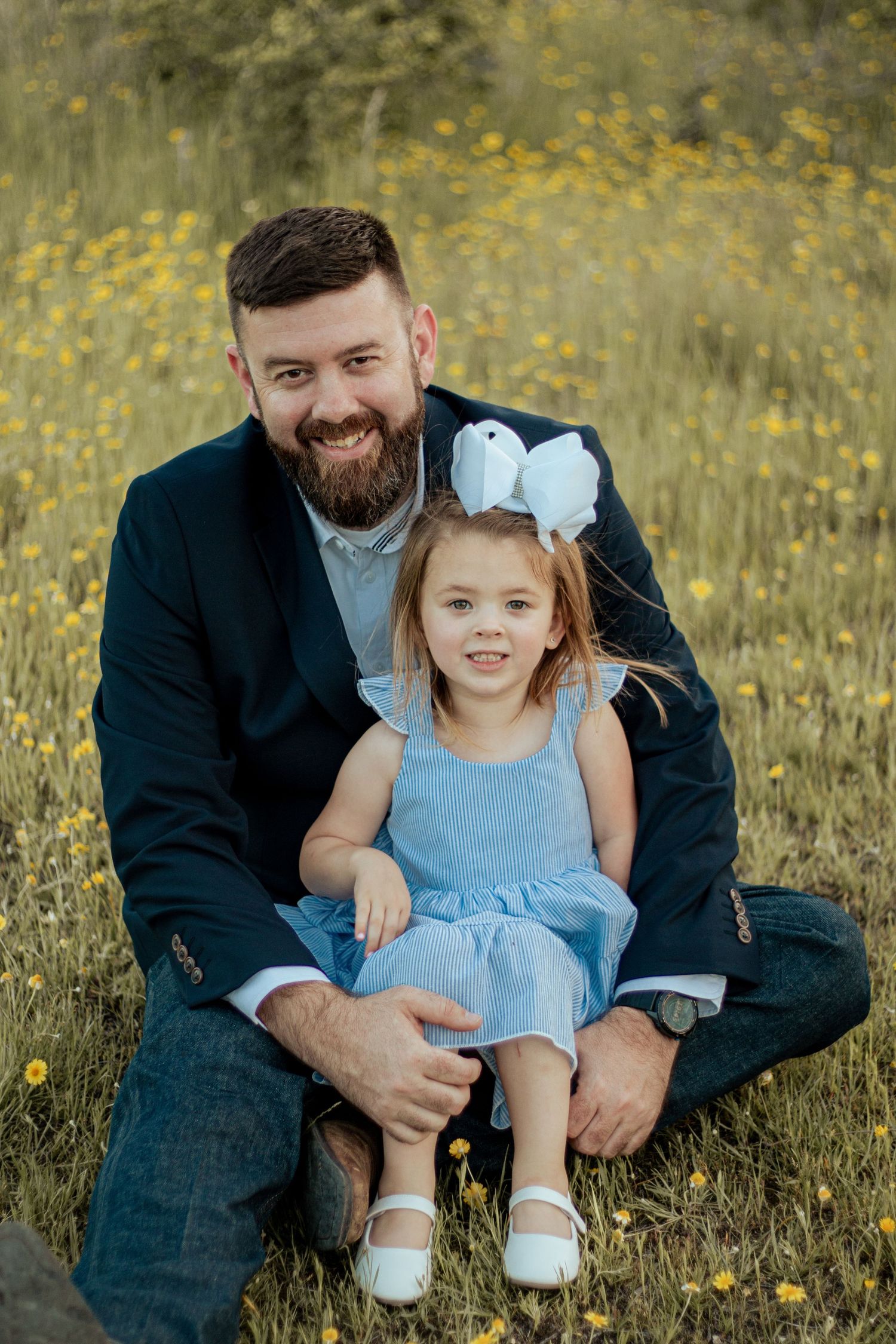 A parent and young child in a blue dress sit together in a field of yellow wildflowers at sunset.