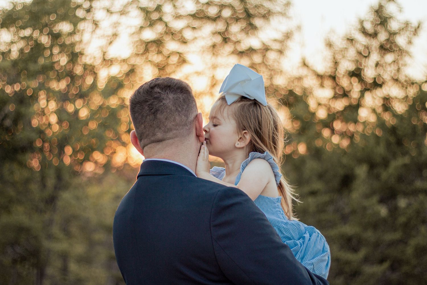 A parent holds a young child in blue dress against golden sunset light filtering through trees.