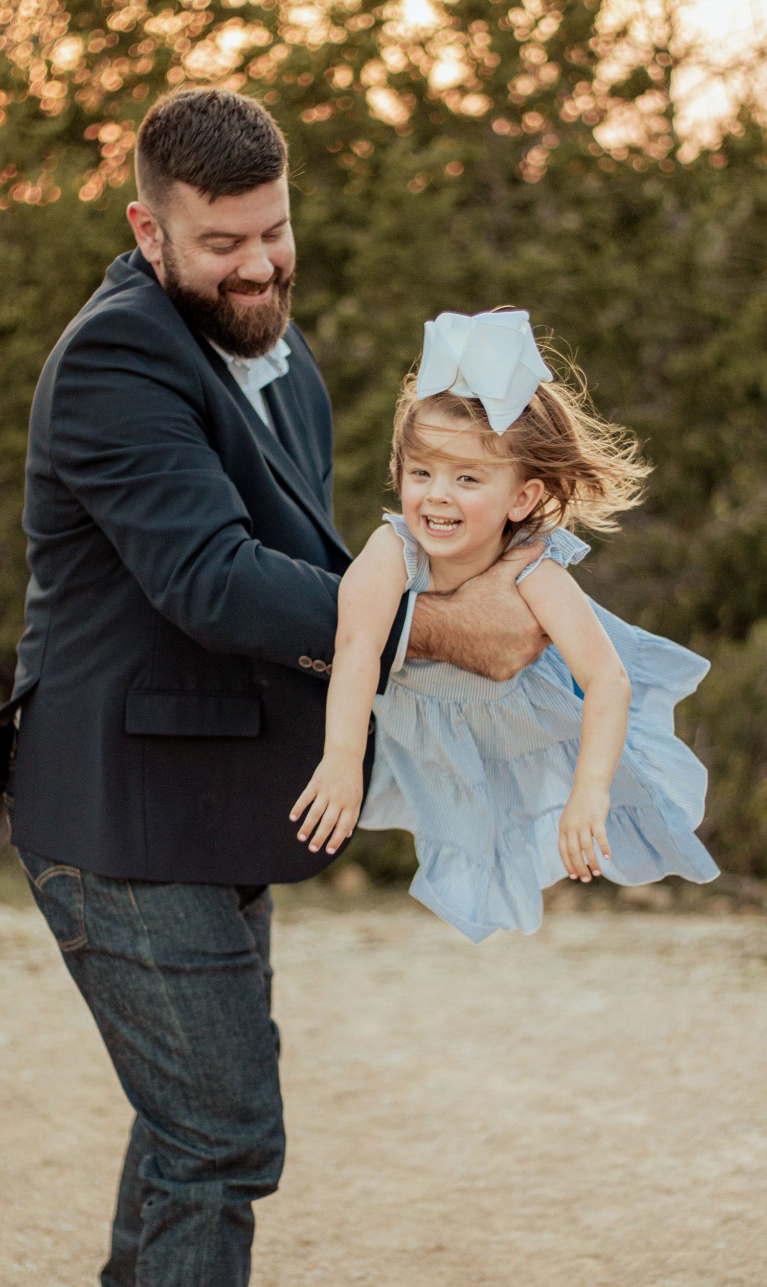 A bearded man in navy blazer playfully lifts a smiling child wearing a light blue dress during golden hour outdoors.