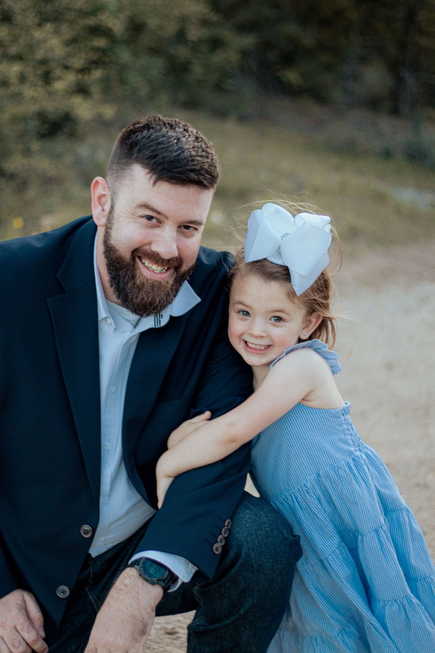 Adult with beard wearing a blue suit kneels beside a small child in a light blue dress and white bow outdoors.