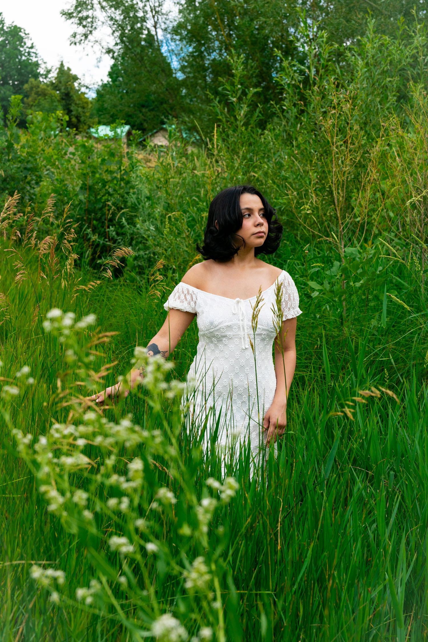 Romantic summer portrait featuring a white off-shoulder dress among tall wild grasses and greenery in nature.