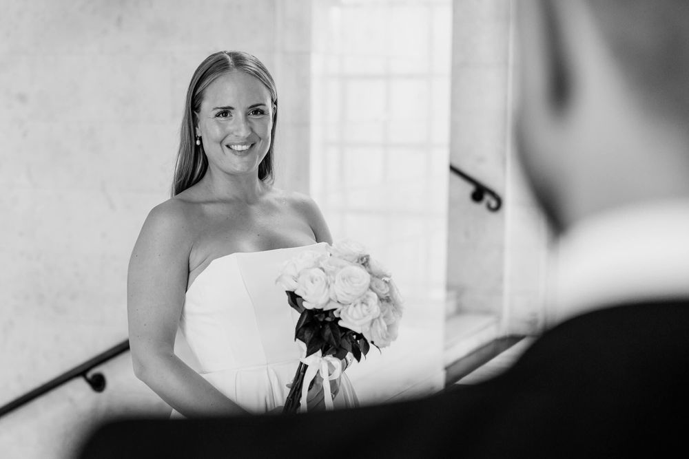 A joyful bride holding a white bouquet stands at the top of a staircase in black and white.
