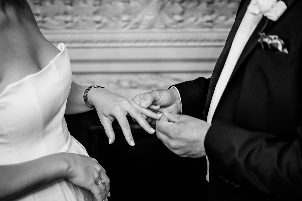 Black and white closeup of exchanging wedding rings during marriage ceremony showing hands and formal attire.