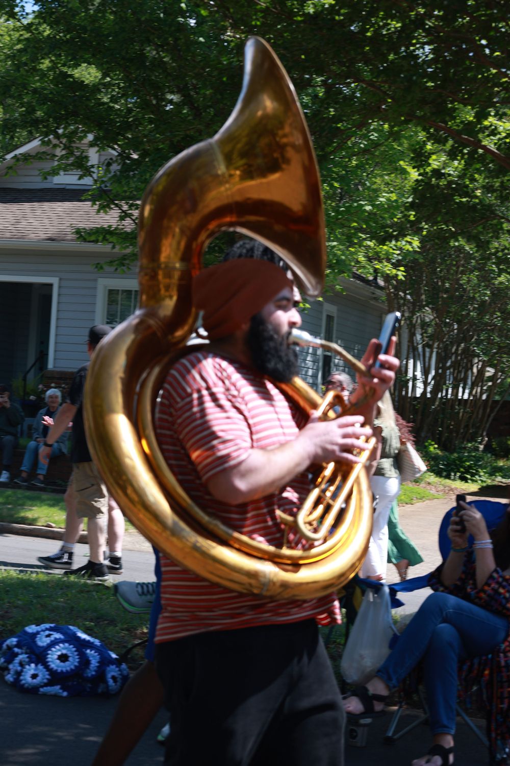 Bearded man playing a large brass sousaphone while holding a phone during an outdoor parade on a sunny day.