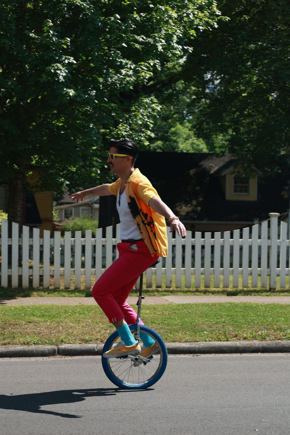 A colorful performer rides a blue unicycle on a street wearing bright pink pants and yellow jacket near a white picket fence.
