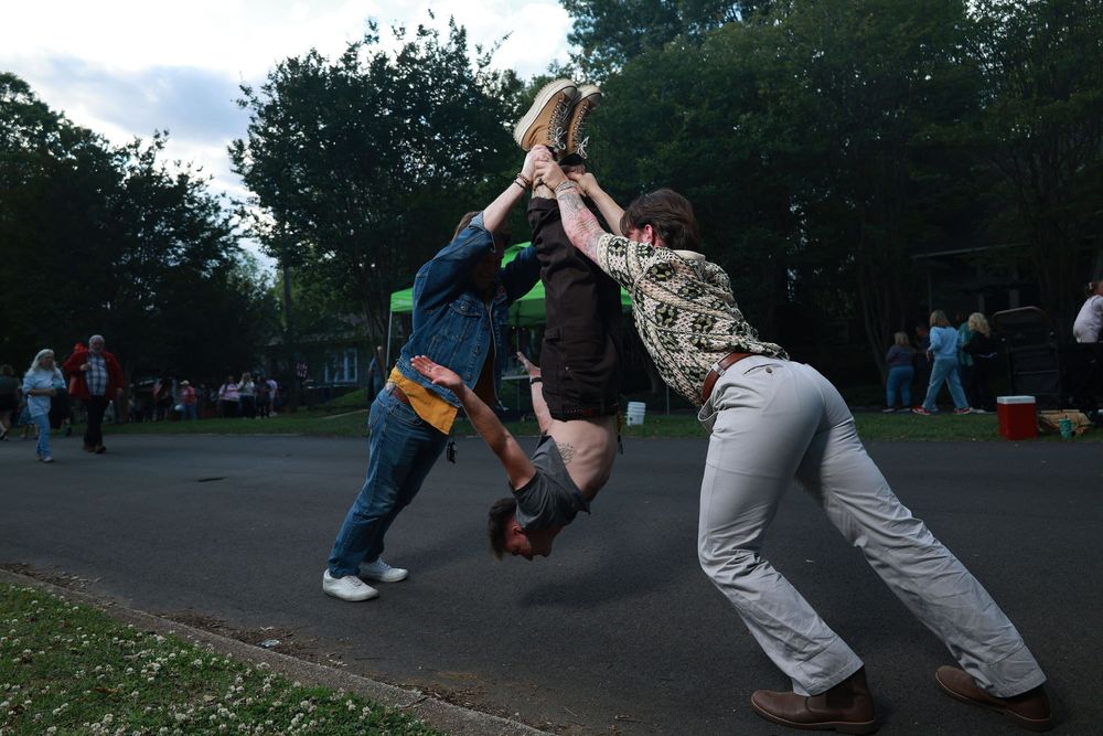 Three people playing an outdoor game in a park, with one person being lifted by two others at dusk.