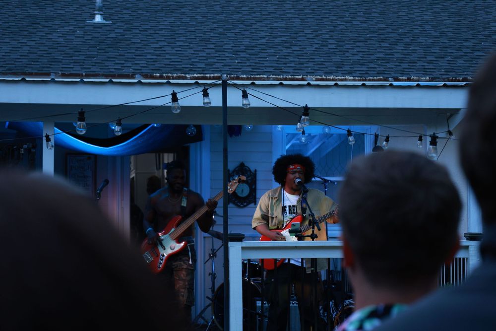 Live band performing outdoors at night, guitarist and vocalist on stage with string lights at a backyard concert.