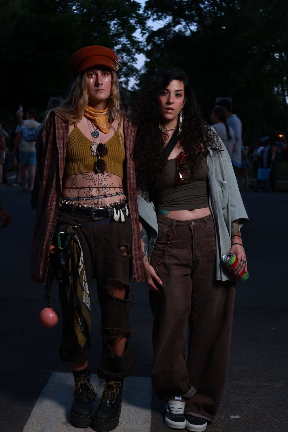 Two festival-goers in bohemian outfits pose at dusk, one in ripped pants and red beret, other in wide-leg trousers.