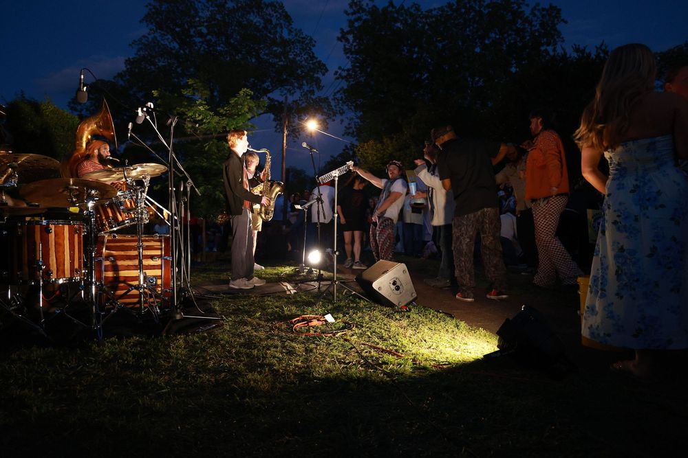Outdoor night concert with band performing on grass stage illuminated by bright light for small crowd.