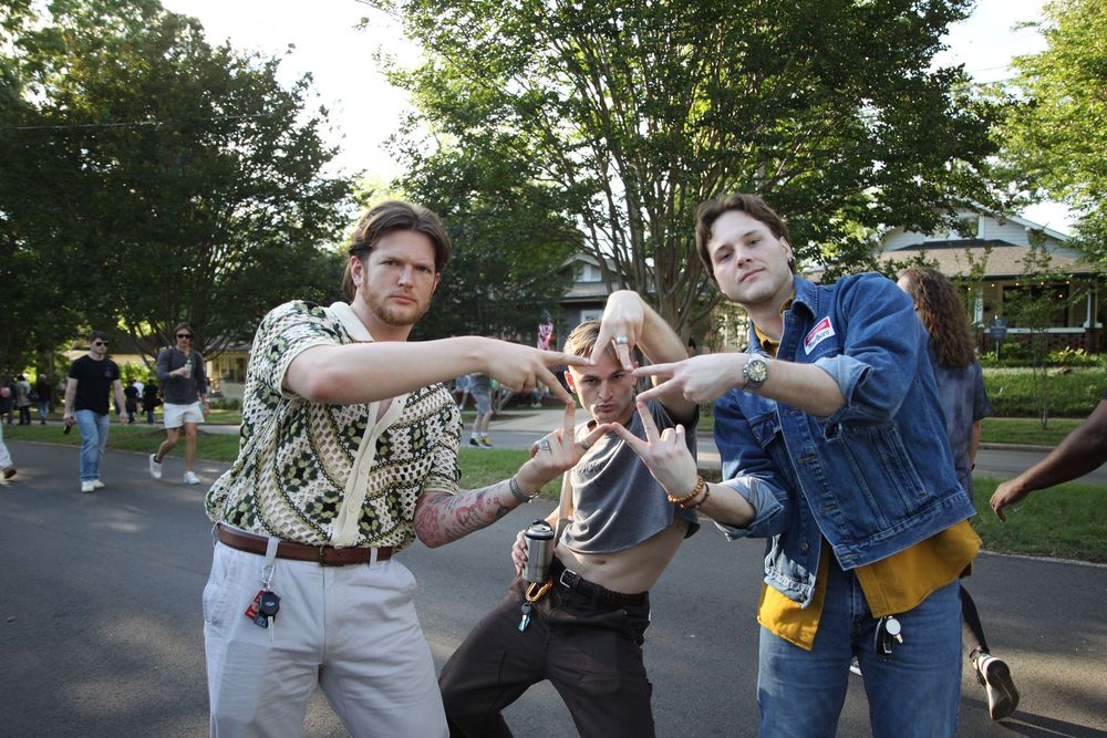 Three young adults posing playfully outdoors at a park event, making fun gestures and smiling at the camera.