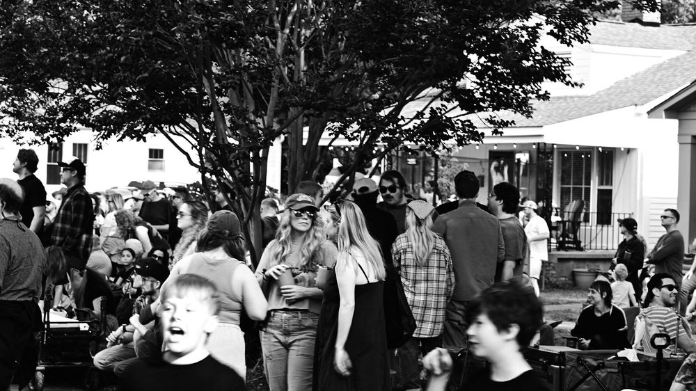 Black and white photo of a lively outdoor gathering with crowd of people socializing near trees and a house.