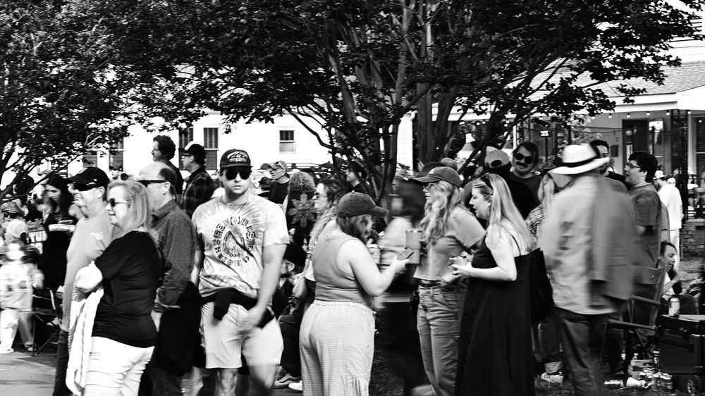 Black and white photo of a large crowd gathered outdoors at a summer festival near trees and buildings.
