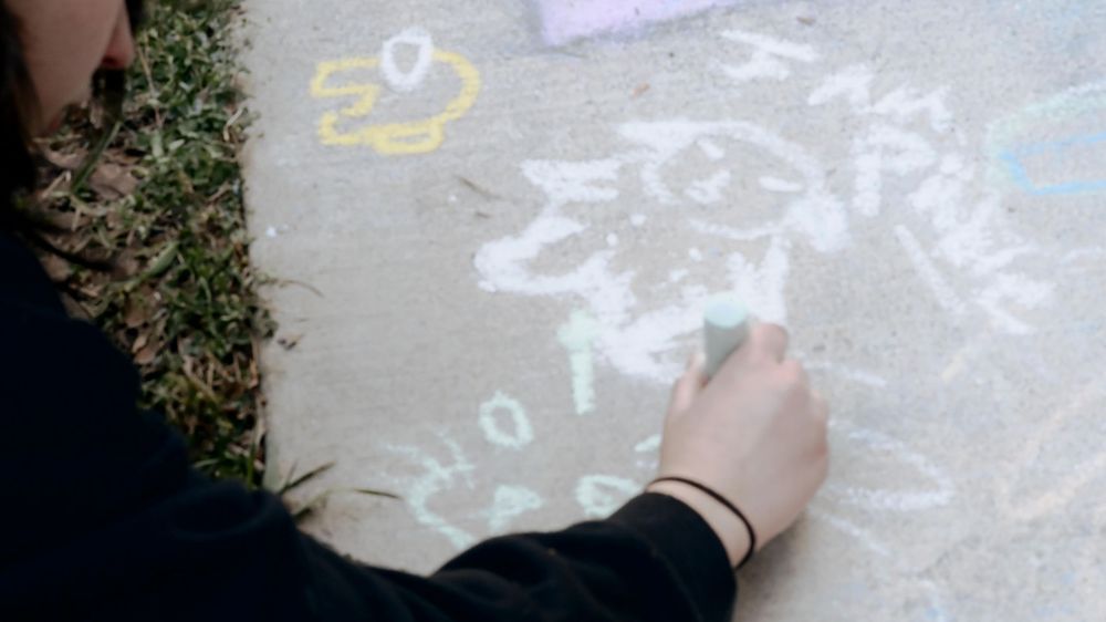 Child drawing colorful chalk art on a sidewalk pavement with letters and shapes visible.
