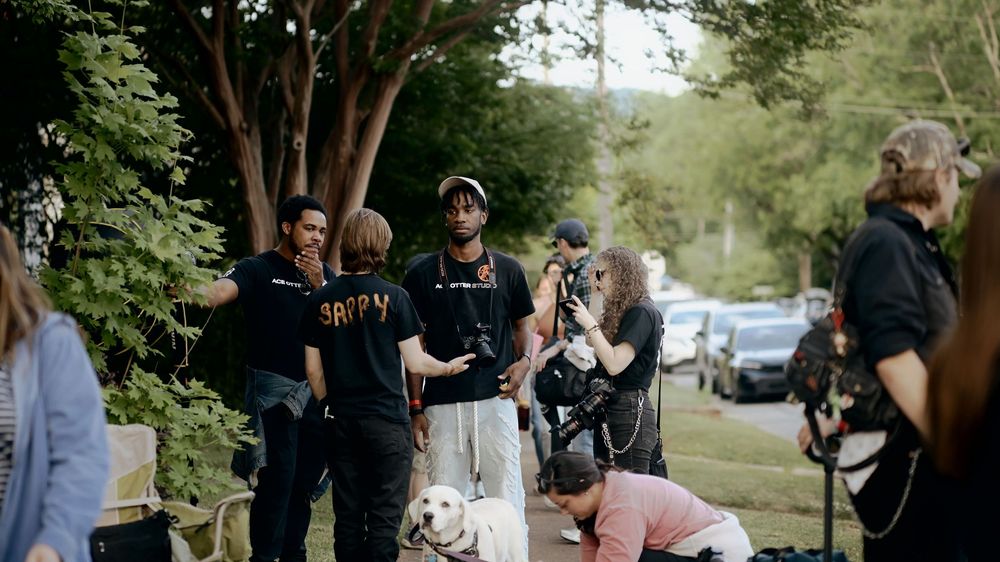 Group of people gathered outdoors on a tree-lined street with a white dog during a casual film shoot.