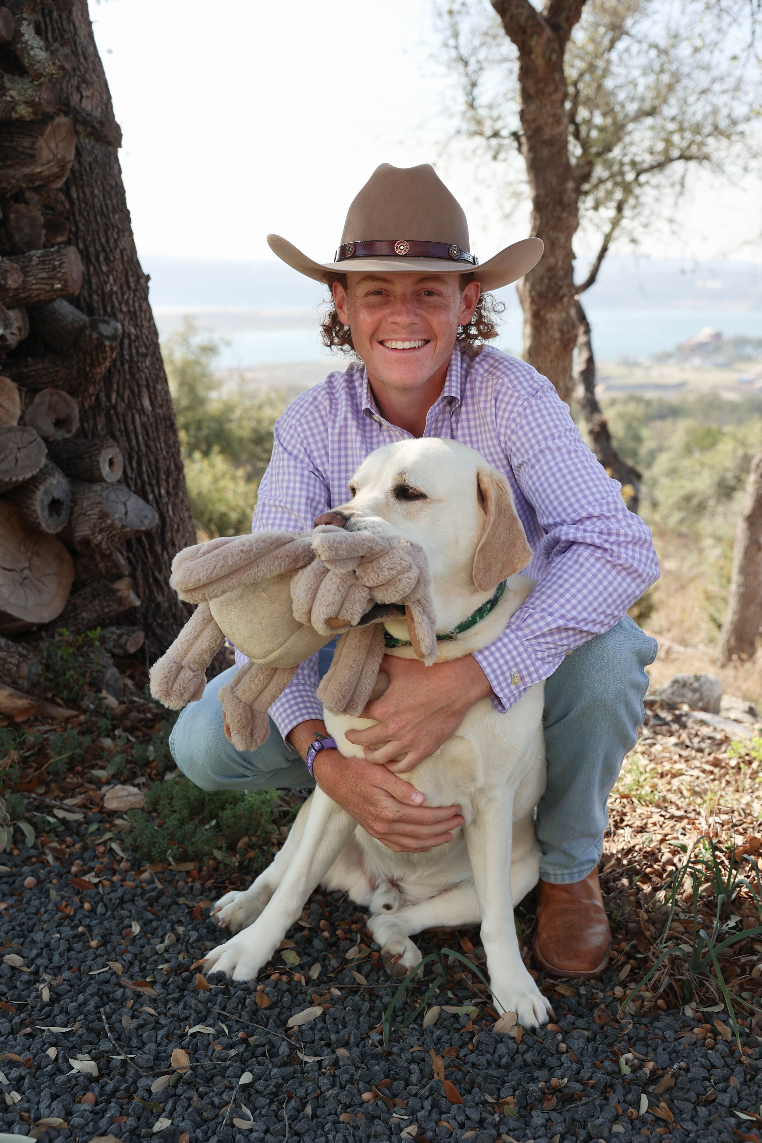 A happy yellow Labrador retriever gets affectionate hugs while sitting outdoors with someone wearing western attire.