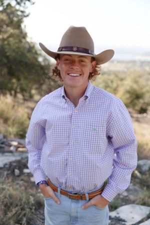 A person wearing a brown cowboy hat and checkered purple shirt poses outdoors in a natural landscape.