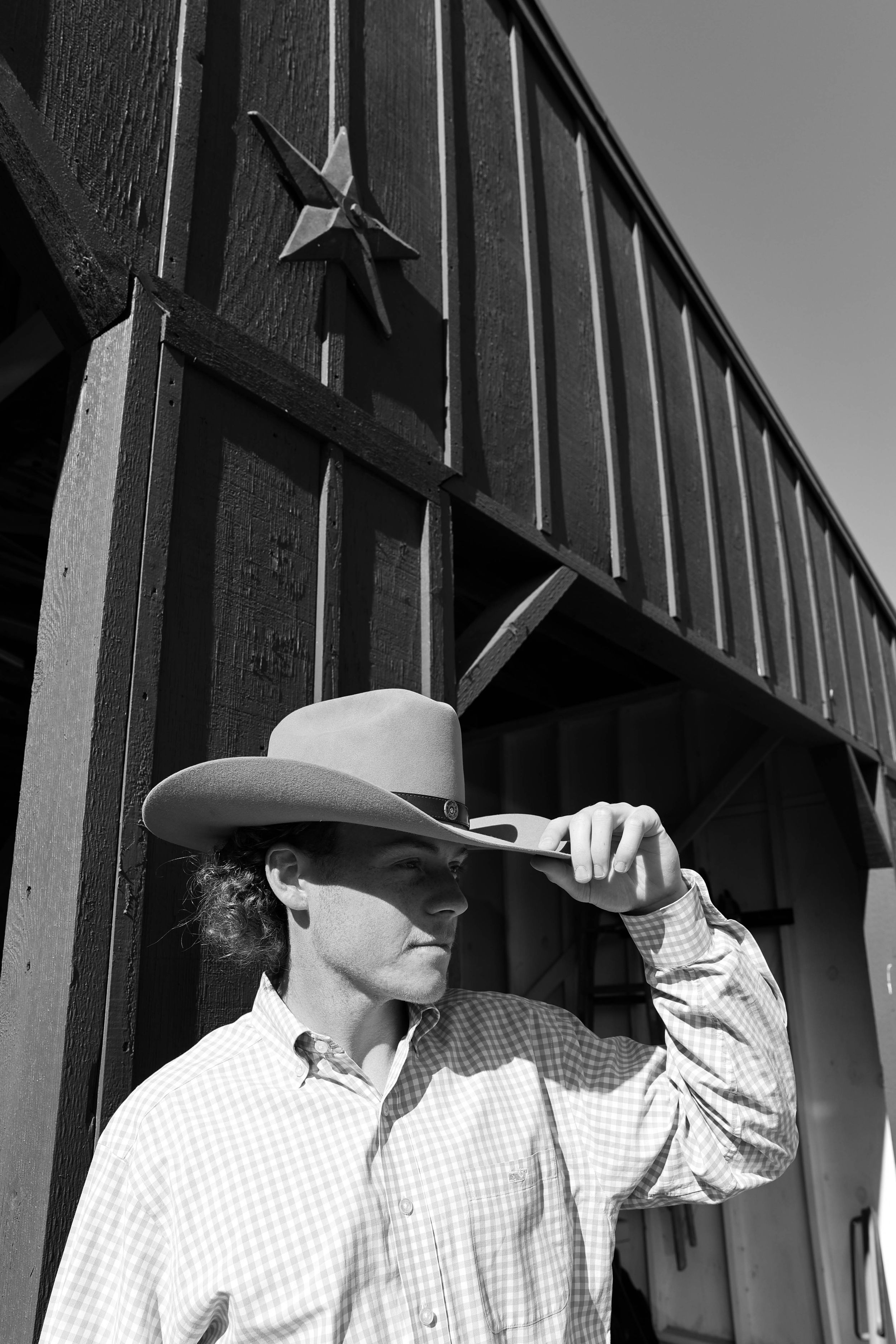 Black and white image of a cowboy in western attire tipping their hat brim against rustic barn siding.