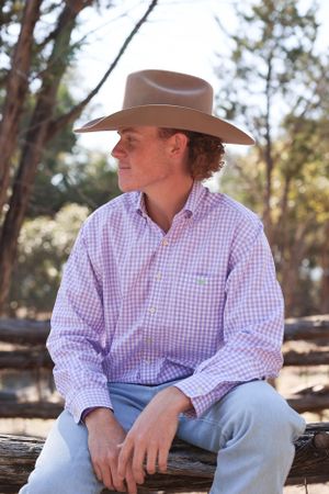 Person in gingham button-down shirt and brown cowboy hat sits pensively on wooden log in outdoor setting.