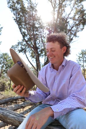 Person in plaid button-down shirt sits on fallen log in sunlit forest while looking at cowboy hat.