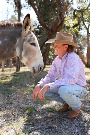 A donkey stands close to someone wearing a cowboy hat and checkered shirt crouching outdoors in a natural setting.
