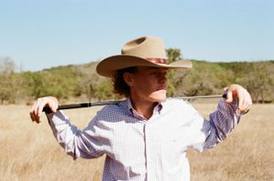 Person in a cowboy hat and checkered shirt stands in a golden grass field holding a pole across their shoulders.