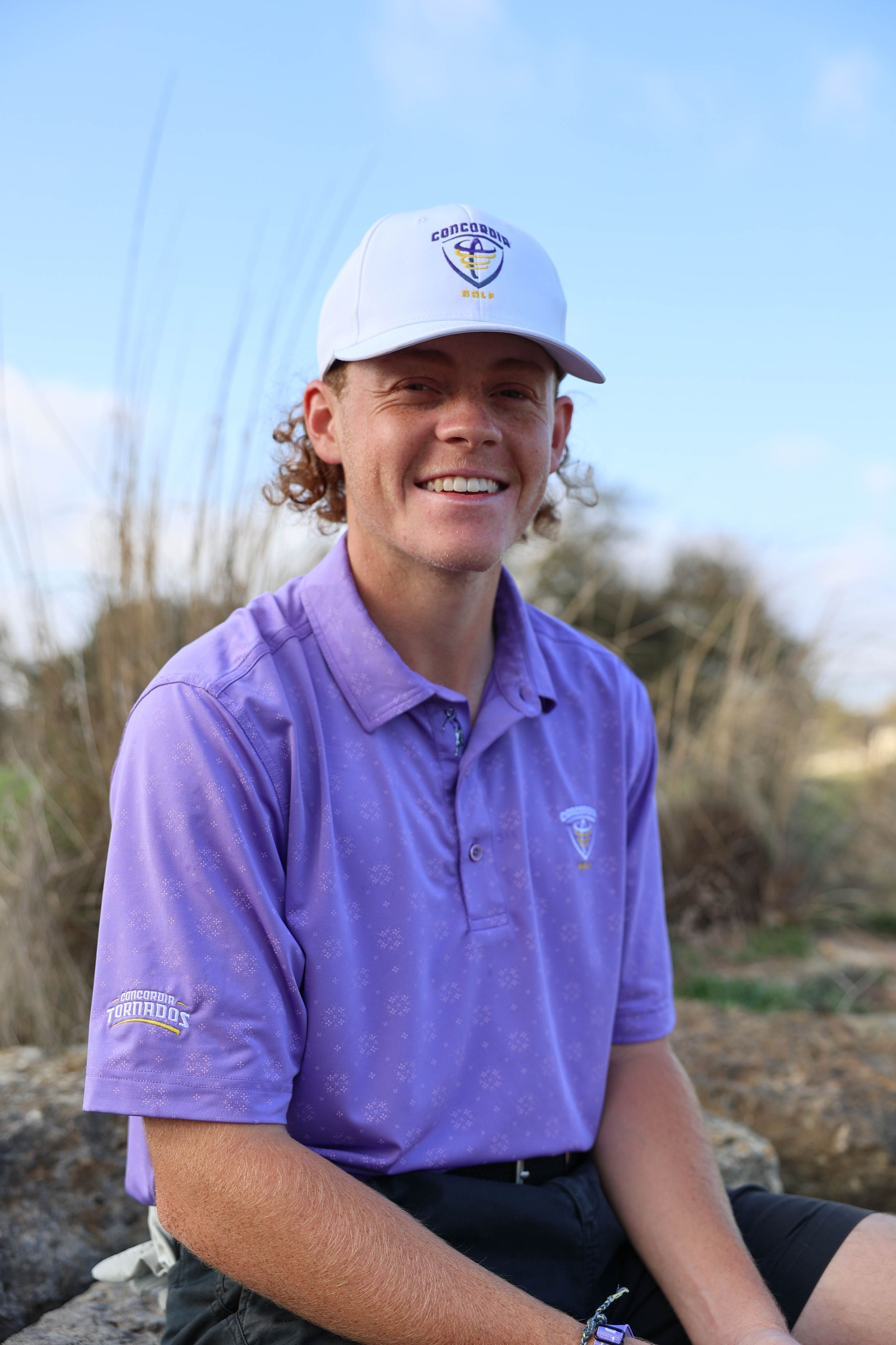 Someone wearing a purple golf polo shirt and white baseball cap sits outdoors on a sunny day.