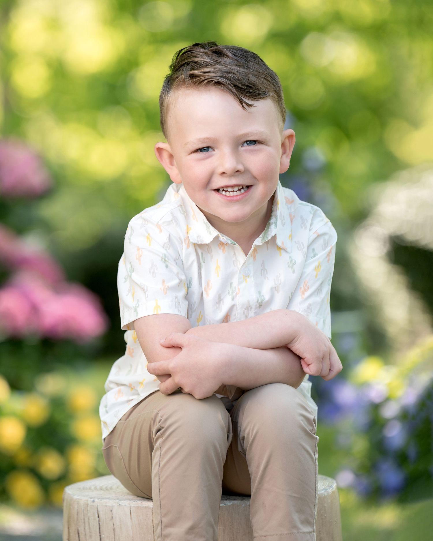 A child in a blue plaid button-up shirt sits on a wooden stump surrounded by colorful garden flowers in the background.